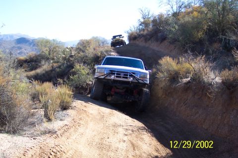 1993 Ford Bronco - Two off-road vehicles navigate a steep, dusty trail in a dry, mountainous landscape.