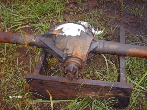 1982 Ford F-100 - A surface-rusted axle housing lies on scrap timber in wet grass with the top of the center section shaved and ground smooth, the bare metal…