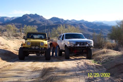 1993 Ford Bronco - A white 1993 Ford Bronco and a yellow Jeep Wrangler are parked on a dirt road in a desert landscape.