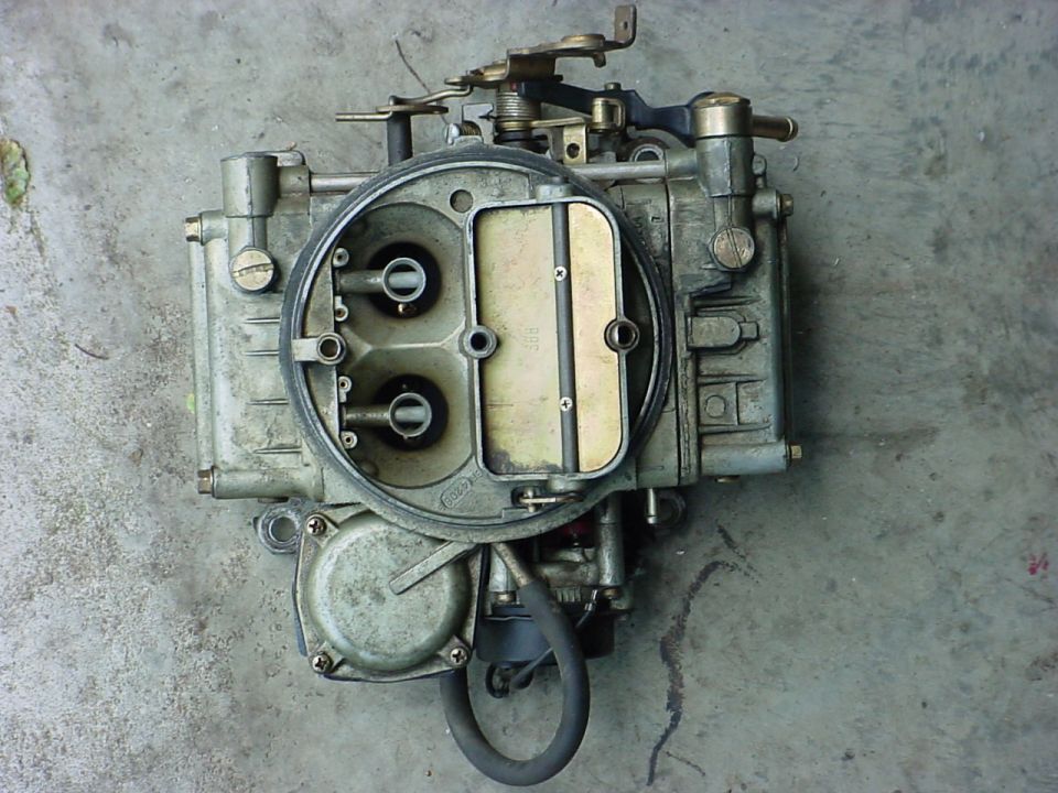Used carburetor with two barrels and metal linkage, resting on a concrete surface