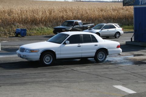1997 Mercury Grand Marquis - A white sedan performs a burnout on a paved surface, with a field of dry crops in the background.