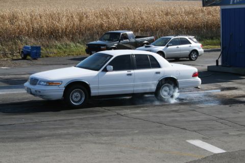 1997 Mercury Grand Marquis - A white sedan performs a burnout on a track, with other vehicles and a cornfield visible in the background.