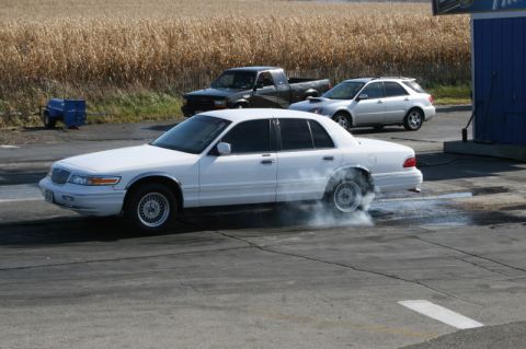 1997 Mercury Grand Marquis - A white 1997 Mercury Grand Marquis performs a burnout at a meet, with a pickup truck and SUV visible in the background.