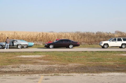 1997 Mercury Grand Marquis - A line of vehicles, including a maroon sedan, a white SUV, and two older cars, are parked on the side of a road with a cornfield behind the…