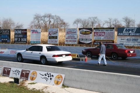 1997 Mercury Grand Marquis - A white 1997 Mercury Grand Marquis and a red sedan are staged on a drag strip, with a man standing between them.