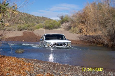 1993 Ford Bronco - This vehicle is driving through a body of water with a dirt bank and vegetation on either side.