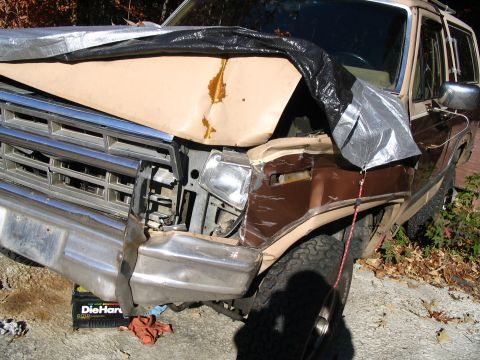 1984 Ford Bronco - SUV with significant front-end damage, partially covered by tarps. A DieHard battery sits below the bumper.