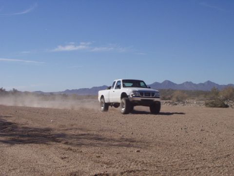 1993 Ford Bronco - A white pickup truck kicks up dust as it drives across a dry, desert landscape under a clear blue sky.