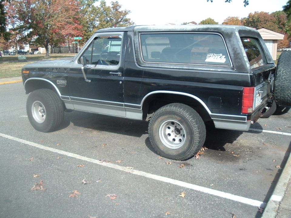 Black Ford Bronco SUV with silver lower trim, side view in a parking lot with fall foliage