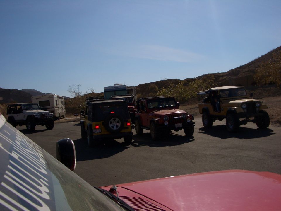 Several Jeeps and RVs parked on a paved area with a hilly, dry landscape under a clear blue sky