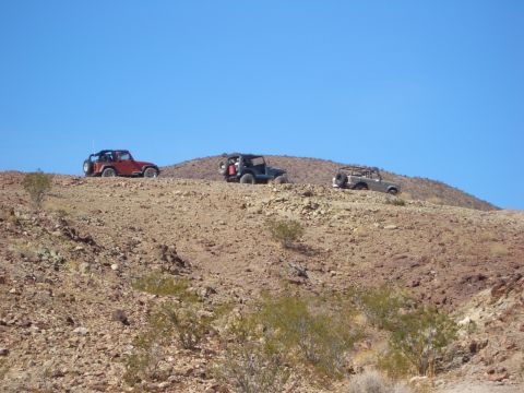 1985 Ford Bronco - Three off-road vehicles traverse a rocky desert landscape under a bright blue sky.