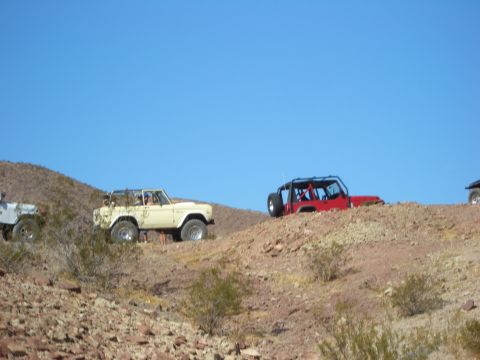 1985 Ford Bronco - Several off-road vehicles navigate a dusty, rocky terrain under a bright blue sky.