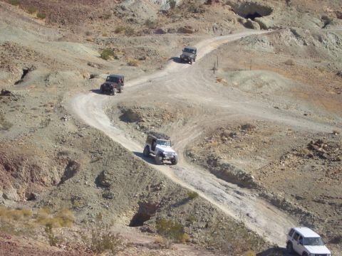 1985 Ford Bronco - Multiple off-road vehicles navigate a dusty, winding trail through a barren, mountainous terrain under a clear sky.