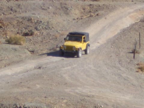 1985 Ford Bronco - A yellow Jeep Wrangler navigates a dusty, unpaved road in a barren, hilly landscape.