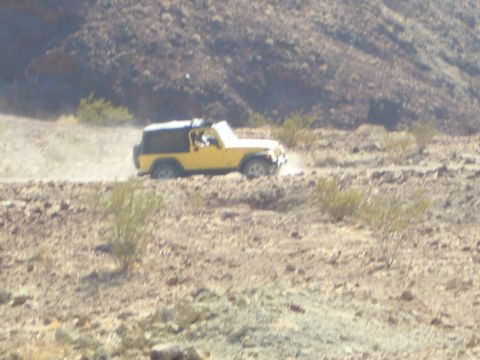 1985 Ford Bronco - A yellow Jeep Wrangler kicks up dust as it travels along a rough, arid terrain.