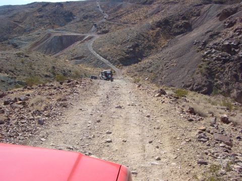 1985 Ford Bronco - Multiple off-road vehicles navigate a winding dirt path through a , mountainous terrain under a clear sky.