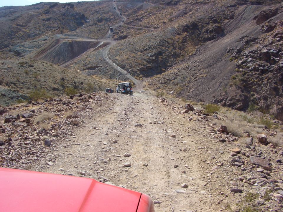 Red vehicle hood in foreground, off-road vehicles on a dirt road in a hilly, arid landscape