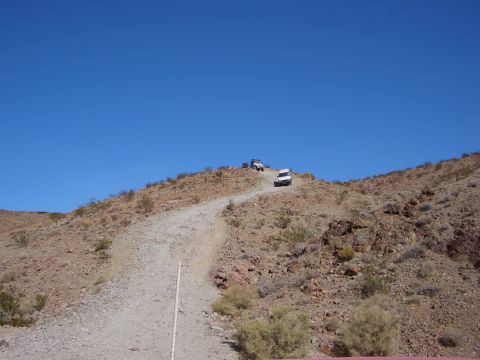 1985 Ford Bronco - Multiple off-road vehicles navigate a challenging dirt path on a desert hillside.