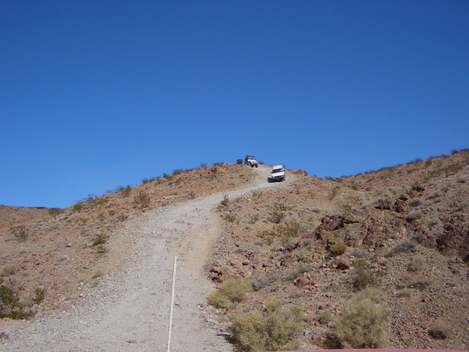 Three off-road vehicles ascend a rocky, dusty hill under a clear blue sky