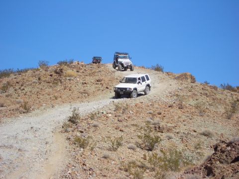1985 Ford Bronco - Several off-road vehicles navigate a dusty, rocky trail on a clear day.