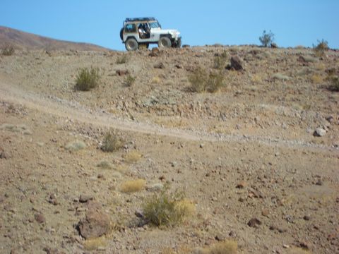 1985 Ford Bronco - A white Jeep Wrangler navigates a dusty, rocky terrain on a clear day.
