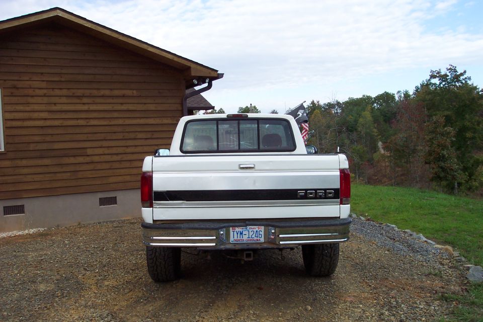 Rear of white Ford pickup with black stripe and NC plate TYM-1246 on gravel driveway by log house