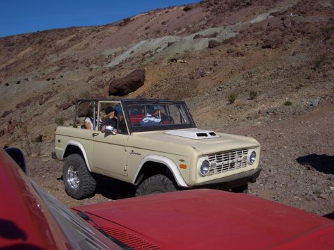 1985 Ford Bronco - A tan Ford Bronco navigates a rocky desert landscape, with another vehicle partially visible.
