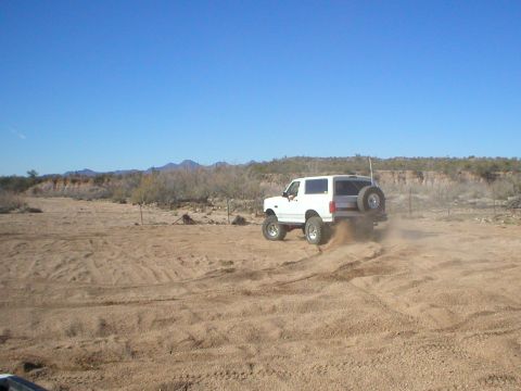 1993 Ford Bronco - White Ford Bronco driving off-road, kicking up dust. Spare tire is visible on the back.