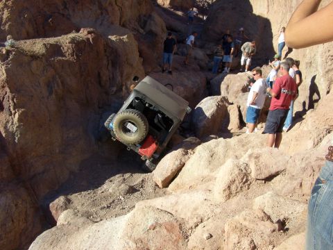 1985 Ford Bronco - A Jeep Wrangler is precariously tilted on a steep, rocky incline as onlookers observe the challenging off-road situation.