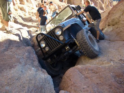 1985 Ford Bronco - A Jeep CJ-7 is carefully driven through a tight, rocky passage with onlookers watching.