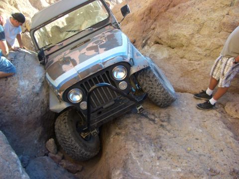 1985 Ford Bronco - A Jeep CJ-7 is wedged between large rocks on an off-road trail, with onlookers observing the situation.
