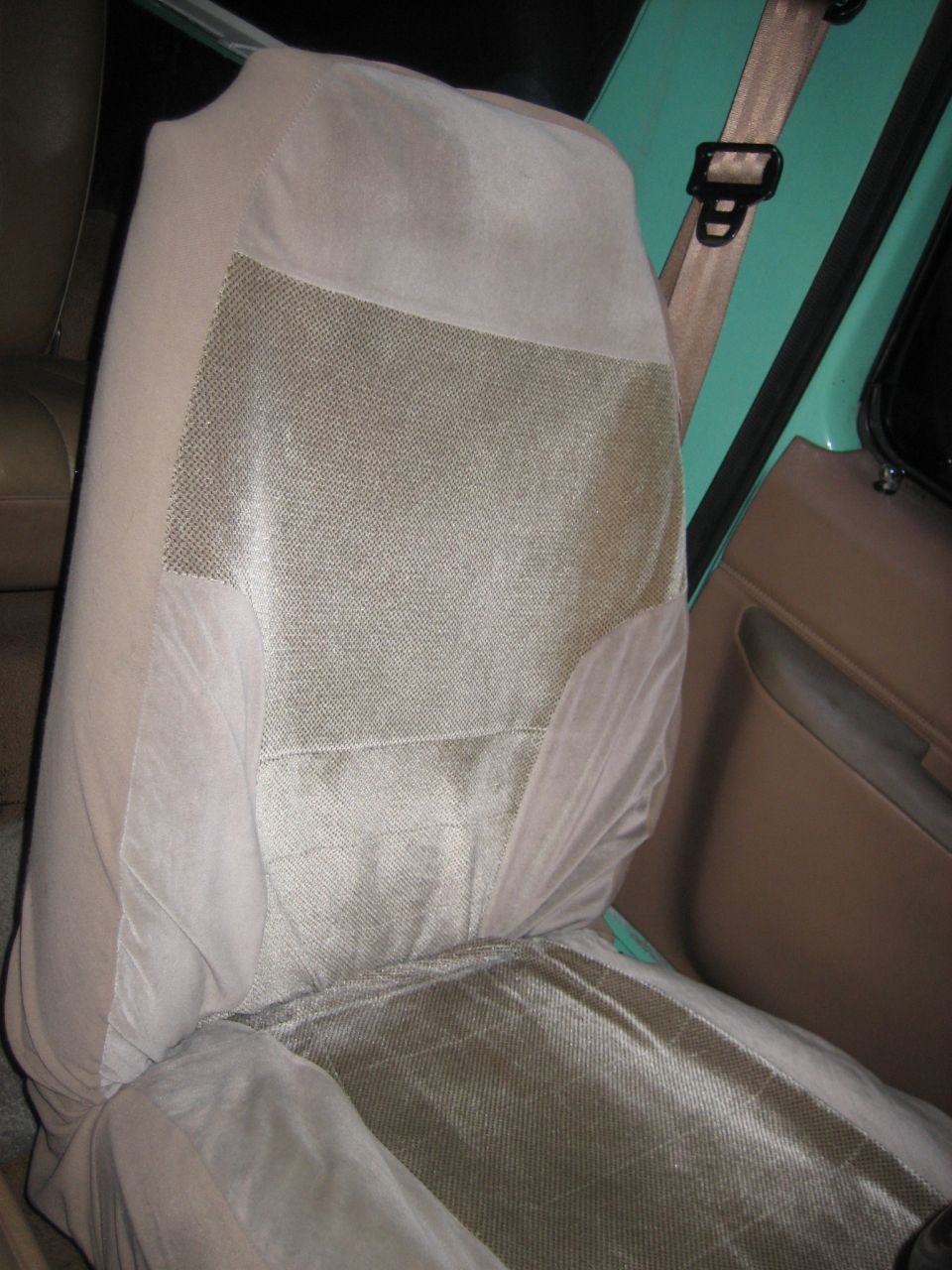 Car seat with a tan seat cover, showing textured fabric on the seat back and cushion. A beige seatbelt is visible