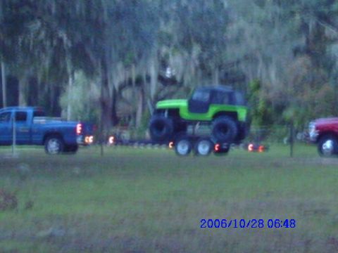 1987 Suzuki Samurai - A blue pickup truck tows a trailer carrying a green off-road vehicle, with another vehicle partially visible on the right.