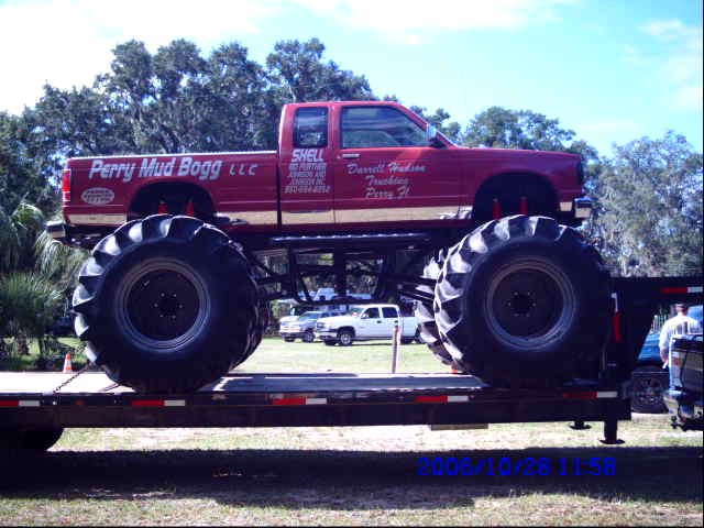Red monster truck with large tires on a flatbed trailer. Text on truck reads 'Perry Mud Bogg LLC' and 'Darrell Hudson Trucking'