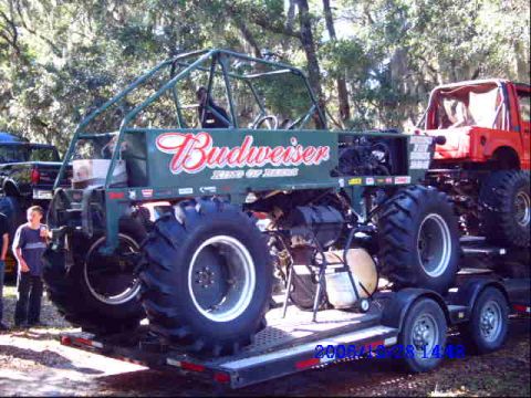 1987 Suzuki Samurai - A custom monster truck with Budweiser livery is parked on a trailer, with a red vehicle visible behind it.
