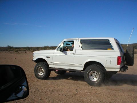 1993 Ford Bronco - A white 1993 Ford Bronco kicks up dust as it drives on a dirt road under a clear blue sky.