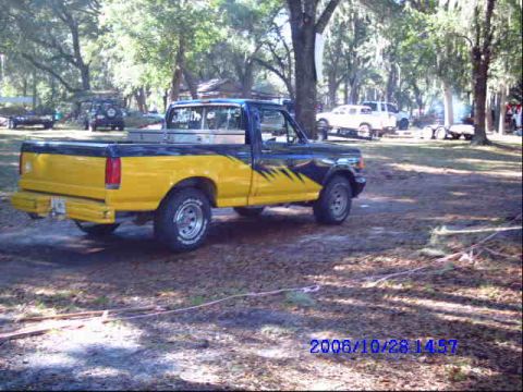 1987 Suzuki Samurai - A two-tone pickup truck sits in a grassy, tree-filled area with other vehicles in the distance.