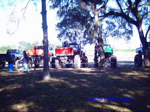 1987 Suzuki Samurai - Several monster trucks are parked outdoors under large trees on a sunny day.