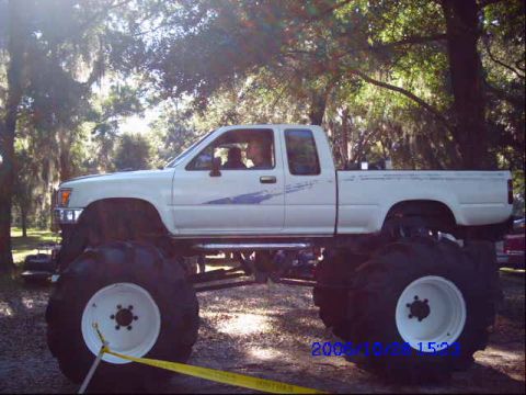 1987 Suzuki Samurai - A modified white pickup truck sits on massive tires under a canopy of trees.