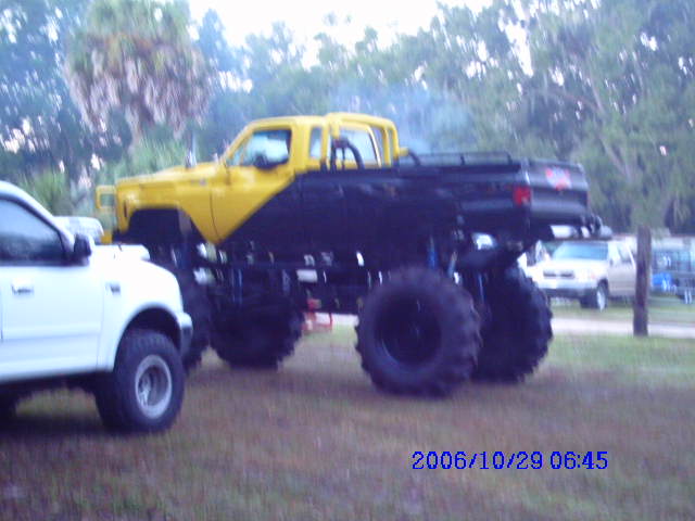 Monster truck with yellow cab and black bed, lifted suspension, large tires. White pickup truck to the left. Trees and grass in background