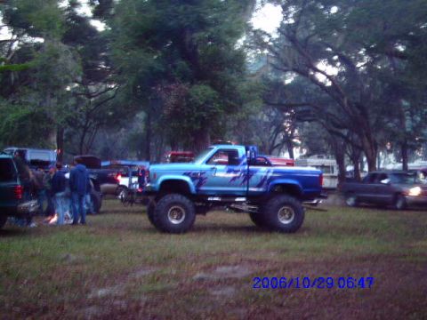 1987 Suzuki Samurai - A lifted blue pickup truck with custom paint sits in a grassy area with trees and other vehicles around.