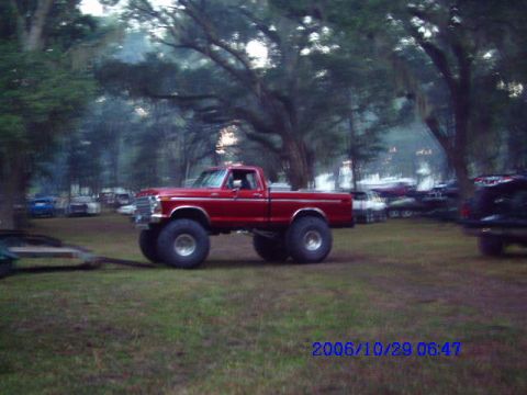 1987 Suzuki Samurai - A red pickup truck with massive tires is parked outdoors among trees and behind other vehicles.