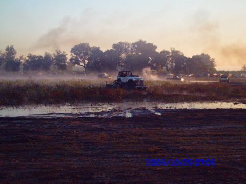 1987 Suzuki Samurai - Multiple off-road vehicles navigate a muddy terrain during an event, with trees and smoke visible in the distance.