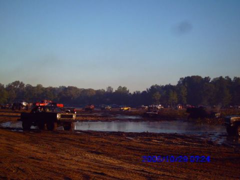 1987 Suzuki Samurai - Multiple trucks are seen navigating a muddy terrain under a clear blue sky, with trees in the background.