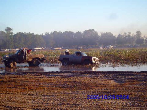 1987 Suzuki Samurai - Two trucks are stuck in a muddy field with vegetation, under a clear sky.