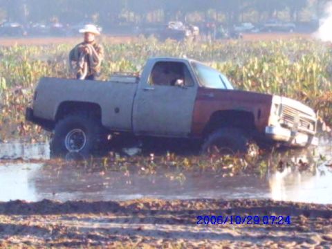 1987 Suzuki Samurai - Pickup truck stuck in mud, with a person in the bed. Grille indicates an early 1980s model.