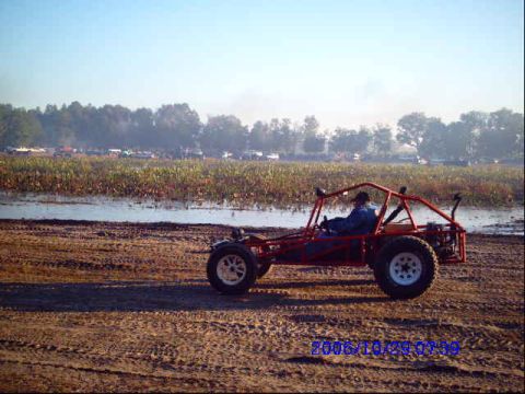 1987 Suzuki Samurai - A person drives a red dune buggy across a dirt track with a body of water and fields in the background.