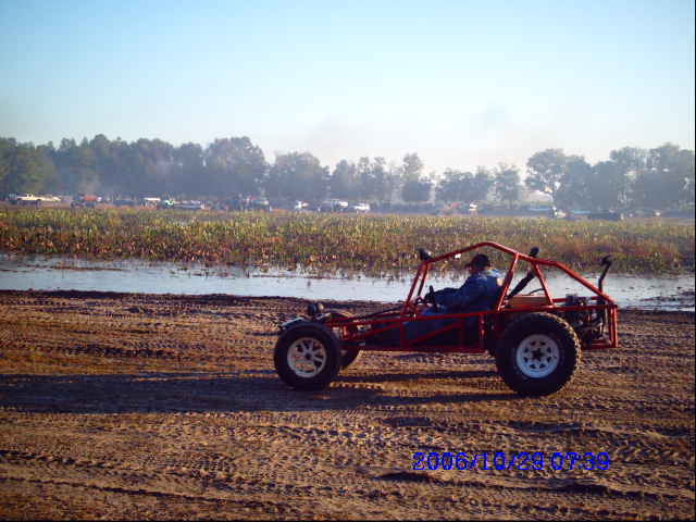 Red dune buggy with driver on dirt track near water and fields