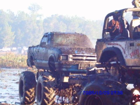 1987 Suzuki Samurai - Two heavily mud-covered vehicles are in a muddy field with trees in the background.