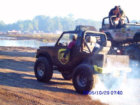 1987 Suzuki Samurai - A brown off-road vehicle with a UPS logo drives through dirt, kicking up smoke, with another vehicle in the background.